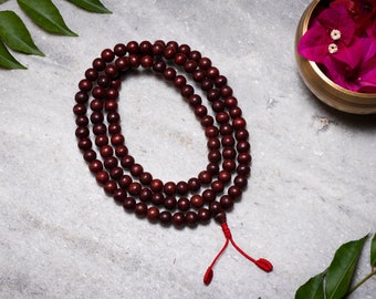 May include: A dark brown wooden mala bead necklace with a red string tassel. The mala beads are arranged in a circle on a light gray surface.