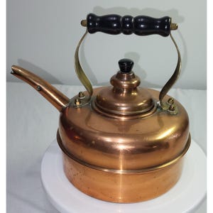 May include: A polished copper tea kettle featuring a rounded body, a long curved spout, and a removable lid with a small black knob. The kettle includes a sturdy metal handle with a central black wooden grip, secured by brass-toned rivets.