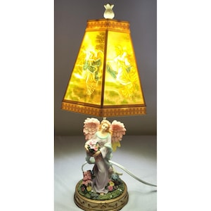 May include: A decorative table lamp featuring an angel figurine base holding a basket of flowers. The lamp has a yellow, trapezoidal shade with a painted scene of figures. The base is surrounded by flowers and foliage.