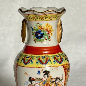 May include: A small, ornate ceramic vase with a white base, featuring a red band and gold accents. The vase is decorated with a colourful painting of figures in traditional clothing. The top and bottom rims have intricate patterns.