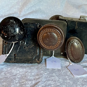 Peut inclure: Un assortiment de quincaillerie de porte ancienne, comprenant un bouton de porte rond noir, un bouton circulaire marron avec un bord décoratif et une plaque ovale marron. La quincaillerie est vieillie avec des signes d'usure visibles.