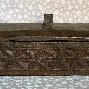 May include: A weathered, rectangular wooden box with a hinged lid and a decorative geometric pattern carved into the front. The box is a dark brown color and has a metal clasp on the side. The box measures approximately 12 inches long.