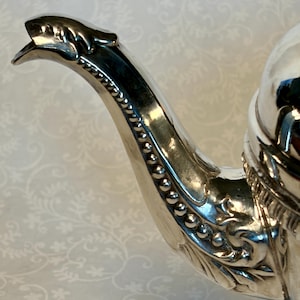 Puede incluir: Primer plano de un pico de tetera plateada con detalles ornamentados. El pico se curva elegantemente, terminando en una cabeza de animal estilizada. El asa está decorada con un borde de cuentas y patrones florales, reflejando un diseño clásico.