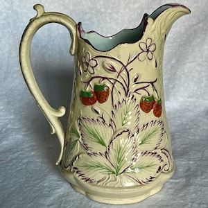 Op de afbeelding: Een vintage keramische kan met een crèmekleurige glazuur en een reliëfdesign van aardbeien en bladeren. De kan heeft een gegolfde rand en een handvat met een rolontwerp.