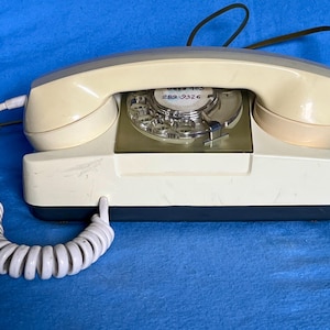 Puede incluir: Teléfono de disco rotatorio crema y blanco con un cable de auricular en espiral. El teléfono tiene una base rectangular con una parte inferior azul oscuro y una cuna de auricular color crema. El dial tiene el número 289-9326 visible. El teléfono está sobre una superficie azul.