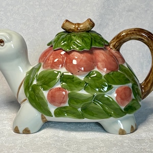 Puede incluir: Tetera de cerámica con forma de tortuga. El caparazón está decorado con manzanas rojas y hojas verdes. El asa y el pico son marrones. La cabeza y las patas de la tortuga son blancas.