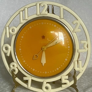 May include: Cream-coloured vintage clock with a mustard-yellow face and white numerals. The clock has a star emblem and a decorative gold-toned stand. The clock's hands are white and indicate the time.