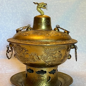 Puede incluir: Olla caliente de latón antiguo con motivos de dragones y nubes. La olla tiene una tapa con un adorno de dragón y asas. La base tiene una placa decorativa. Una pieza única para servir comida.