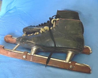 May include: A pair of vintage ice skates. The skates are black leather with metal blades and brown leather straps. The laces are a faded yellow colour. The skates show signs of wear and age, suggesting they are antique or vintage.