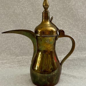Puede incluir: Una cafetera de latón antigua con un pico largo y un adorno decorativo en la tapa. La cafetera tiene un asa curvada y una tapa con bisagras. El latón tiene una pátina cálida y envejecida, con toques de verde y dorado.