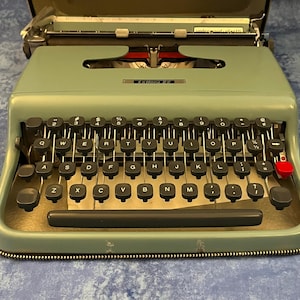 May include: A green vintage typewriter with a black keyboard and the text "Lettera 22" on the front. The typewriter has a red button on the right side.