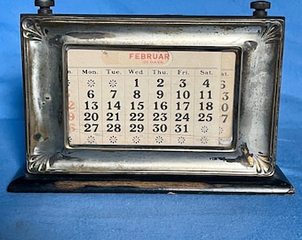 Pode incluir: Um calendário de mesa antigo com uma moldura de metal prateado e uma base preta. O calendário exibe o mês de fevereiro, com os dias impressos em um pequeno cartão de papel. Possui dois pequenos botões na parte superior.