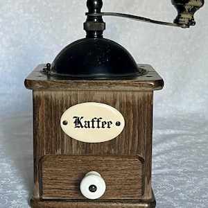 May include: A vintage wooden coffee grinder with a black top and handle. The grinder has a drawer with a white knob and a label that reads "Kaffee". The wood has a natural grain.