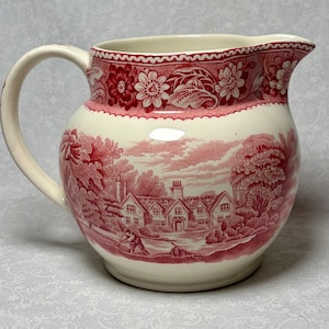 May include: Cream-colored pitcher with a red floral and landscape design. The pitcher features a detailed illustration of a rural scene with houses, trees, and figures. The rim and handle are also cream-colored.