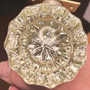 Op de afbeelding: Een heldere, facetgeslepen glazen deurknop met een decoratief, stervormig midden. De knop heeft een geschulpte rand en is gemonteerd op een messingkleurige metalen plaat. Het glas reflecteert licht.