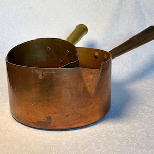 Könnte beinhalten: Ein Kupfertopf mit zwei Abschnitten und zwei Messinggriffen. Der Topf hat eine abgerundete Form mit einem Ausguss auf einer Seite. Das Kupfer hat eine warme, rotbraune Farbe, und die Griffe sind goldfarben. Dieses Vintage-Kochgeschirr ist ideal zum Kochen.