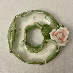 May include: A ceramic, ring-shaped planter in green and white. It features a detailed rose with pink petals and green leaves. The design includes a central opening and a textured, leaf-like border. The planter is likely for decorative use.