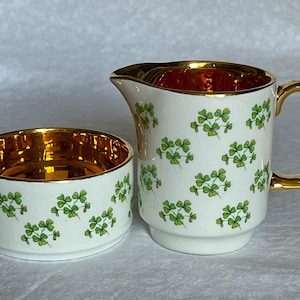 Pode incluir: Um conjunto de cremeira e açucareiro em cerâmica branca. O exterior é decorado com trevos verdes. O interior e a pega da cremeira são dourados. O açucareiro é redondo e a cremeira tem pega e bico.