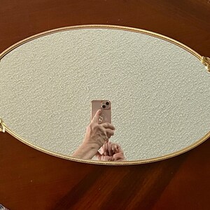 May include: Oval gold-toned mirror with leaf detailing at each end. The mirror reflects a person holding a phone. The mirror is set on a dark brown wooden surface.