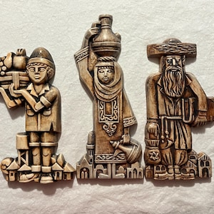 Può includere: Tre placche decorative da parete che raffigurano figure in abiti tradizionali. Le figure sono scolpite in un colore marrone chiaro e presentano dettagli elaborati. Le placche sono montate su uno sfondo bianco.