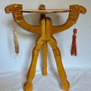 May include: A light brown wooden folding table featuring three legs carved into the shape of camel heads at the top. The table has a circular top surface and includes two decorative hanging tassels, one beige and one reddish-orange.