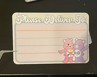 May include: A white card with the text "Please Deliver To:" in a retro font. There are five lines for writing an address. The card features a pink and purple cartoon bear with a rainbow heart on a life preserver.