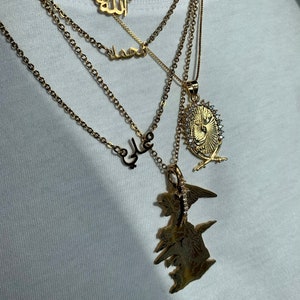 May include: A gold chain necklace with four pendants. The pendants are all in Arabic script. The top pendant says "Allah", the second pendant says "Muhammad", the third pendant says "Ali", and the bottom pendant is a large, ornate design with a crown and swords.