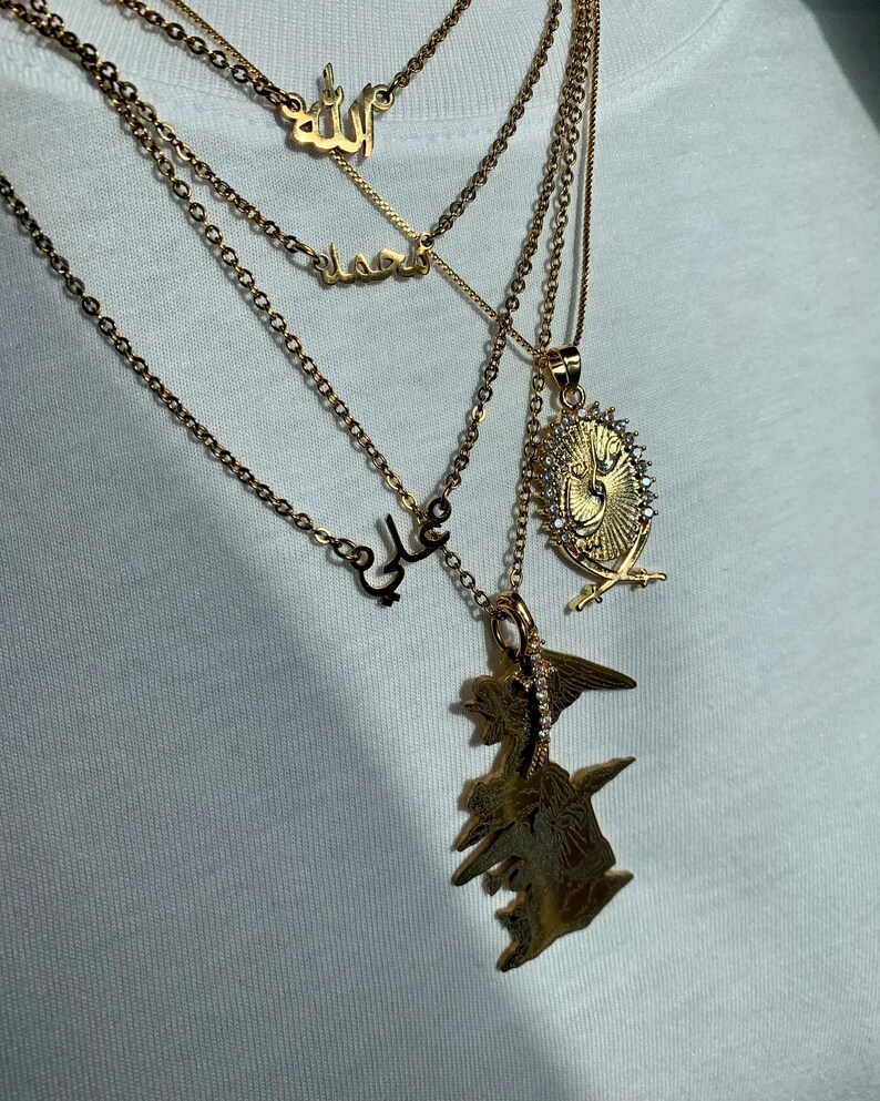 May include: A gold chain necklace with four pendants. The pendants are all in Arabic script. The top pendant says "Allah", the second pendant says "Muhammad", the third pendant says "Ali", and the bottom pendant is a large, ornate design with a crown and swords.
