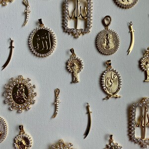 May include: A collection of gold pendants with various designs, including swords, trees, and Arabic calligraphy. The pendants are adorned with sparkling clear crystals.