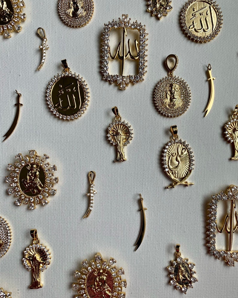 May include: A collection of gold pendants with various designs, including swords, trees, and Arabic calligraphy. The pendants are adorned with sparkling clear crystals.