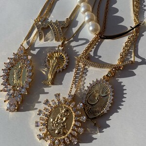 May include: A set of gold necklaces with various pendants, including a religious figure, a stylized Arabic word, and a tree. The necklaces are adorned with clear crystals and are layered on a white surface.