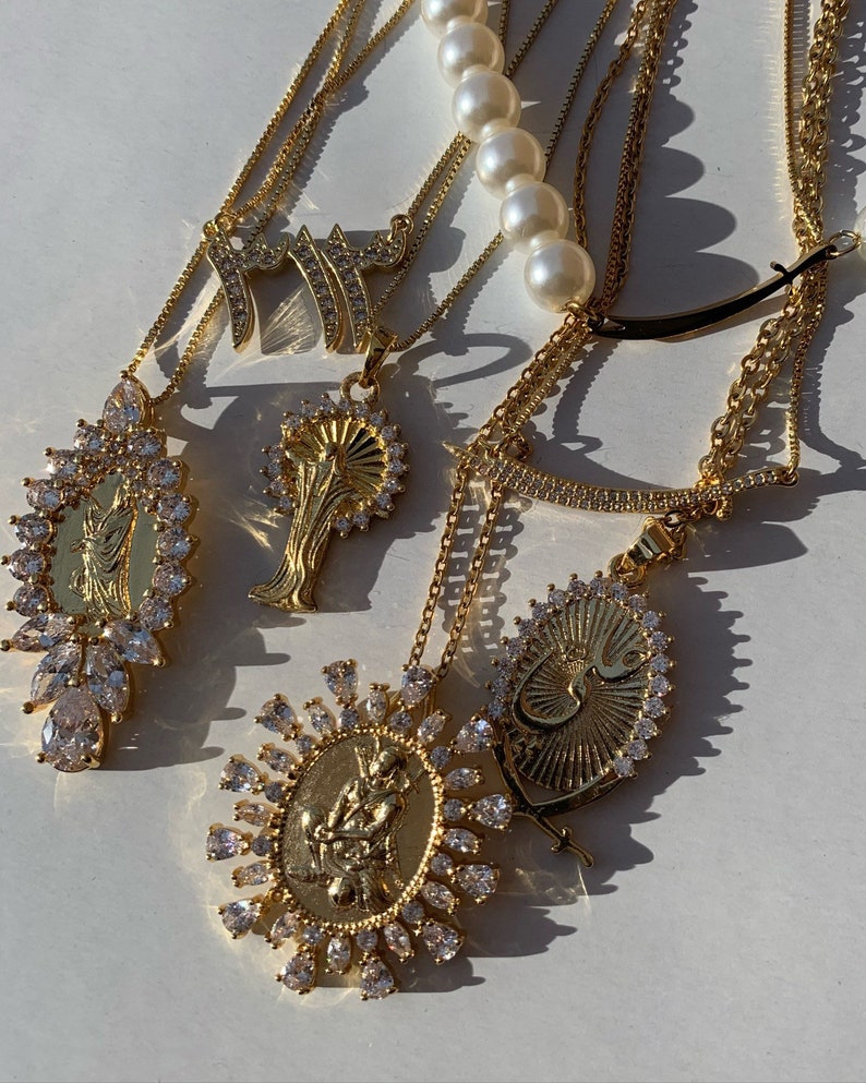 May include: A set of gold necklaces with various pendants, including a religious figure, a stylized Arabic word, and a tree. The necklaces are adorned with clear crystals and are layered on a white surface.