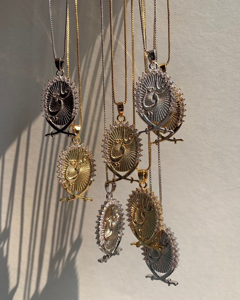May include: A group of seven silver and gold necklaces with pendants featuring a stylized Arabic inscription surrounded by a circle of clear rhinestones. Each pendant is attached to a delicate chain.