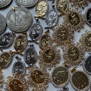 May include: A collection of gold and silver pendants with intricate designs and clear crystals. The pendants feature Arabic calligraphy and religious symbols.