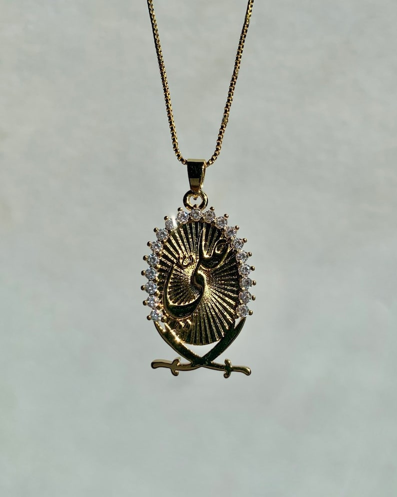 May include: A gold chain necklace with a pendant featuring a gold oval with a sunburst design and a calligraphic symbol surrounded by clear rhinestones. Two crossed swords are below the oval.