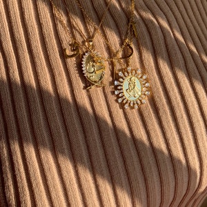May include: A gold chain necklace with multiple pendants, including a heart, a letter, and two oval-shaped pendants with a religious figure and a cross design. The pendants are adorned with sparkling clear rhinestones.