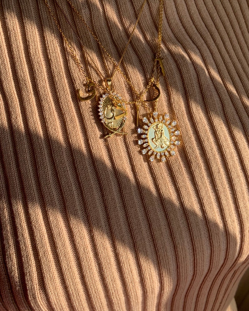 May include: A gold chain necklace with multiple pendants, including a heart, a letter, and two oval-shaped pendants with a religious figure and a cross design. The pendants are adorned with sparkling clear rhinestones.