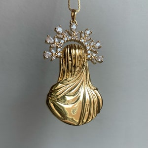 Könnte beinhalten: Goldfarbene Halskette mit Anhänger, die eine religiöse Figur mit drapiertem Schleier und Heiligenschein aus klaren Kristallen zeigt. Der Anhänger hängt an einer zarten Goldkette, ein elegantes und spirituelles Accessoire.
