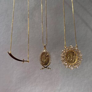 May include: Three gold necklaces with different pendants. The first necklace has a crescent moon and sword pendant. The second necklace has a pendant with Arabic calligraphy and two swords. The third necklace has a pendant with Arabic calligraphy surrounded by clear rhinestones.