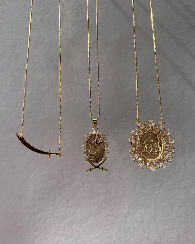 May include: Three gold necklaces with different pendants. The first necklace has a crescent moon and sword pendant. The second necklace has a pendant with Arabic calligraphy and two swords. The third necklace has a pendant with Arabic calligraphy surrounded by clear rhinestones.