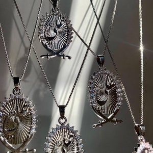May include: Five silver necklaces with oval pendants featuring a stylized Arabic script design surrounded by a circle of small clear stones. Each pendant has two crossed swords at the bottom.
