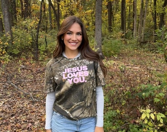 May include: A camouflage-patterned t-shirt with the text "JESUS LOVES YOU" in pink letters. The shirt is paired with light blue jeans and a long-sleeved gray shirt underneath. The background is a forest with trees and fallen leaves.
