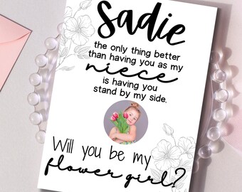 Könnte beinhalten: Eine weiße Karte mit schwarzer Schrift: „Sadie, das einzig Besseres, als dich als meine Nichte zu haben, ist, dich an meiner Seite zu haben.“ und „Willst du mein Blumenmädchen sein?“ Ein Foto eines jungen Mädchens mit Blumen in der Mitte. Ein rosa Umschlag im Hintergrund.