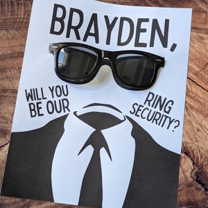 Op de afbeelding: Een witte kaart met zwarte tekst die "Brayden, Will you be our Ring Security?" zegt. Een paar zwarte zonnebrillen ligt op de tekst.