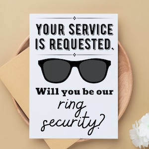 May include: A white card with black text that reads "Your service is requested."  A pair of black sunglasses are above the text. Below the sunglasses is the text "Will you be our ring security?"