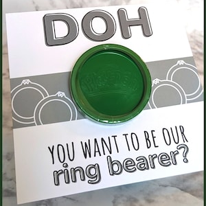 May include: A white card with the word "DOH" in large black letters. Two gray rings are on the left side of the card. A green Play-Doh container is in the center of the card. The card reads "You want to be our ring bearer?"