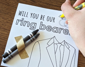 May include: A printable card with the text "Will you be our ring bearer?" with a drawing of a suit and tie, two crayons, and a gold ribbon.