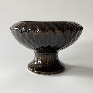 Può includere: Una ciotola con piedistallo in ceramica marrone scuro con esterno strutturato e scanalato. La ciotola ha una parte superiore ampia e aperta e poggia su una base a piedistallo corta e arrotondata. La finitura lucida riflette la luce, evidenziando il design dettagliato.