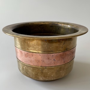 May include: A brass planter with a copper band around the middle. The planter has a wide rim and a rounded bottom.