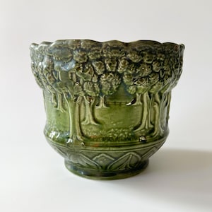May include: A green ceramic planter with a detailed forest design. The planter has a scalloped rim and a pedestal base. The forest scene is embossed around the body. The planter is approximately 15 cm tall.